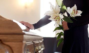 A person in black clothing holds white lilies, placing a hand on a wooden casket in a softly lit room, suggesting a funeral or memorial service.