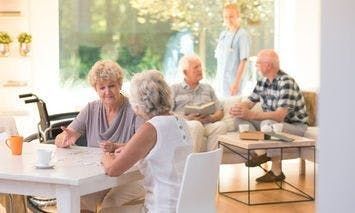 Elderly women sitting at a table converse in a bright room, while a nurse stands, and two elderly men seated on a sofa chat; a wheelchair and coffee cups are visible.