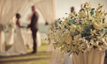 A bouquet of white and blue flowers sits in the foreground of an outdoor wedding ceremony, with a wedding couple standing under a canopy in the blurred background.